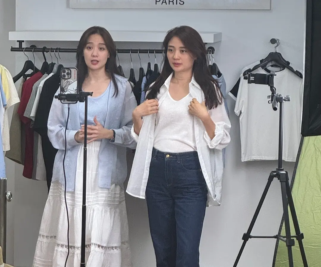 라이브커머스 방송 현장 — 진행자 두 명 앞에 촬영용 스마트폰과 조명 스탠드가 놓여 있고 뒤로 의류 행어가 있습니다.